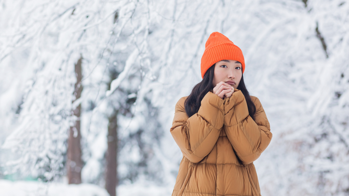 Eine junge Frau mit ernstem Blick wärmt ihre Hände, trägt eine orangefarbene Mütze und Daunenjacke vor verschneiten Bäumen.