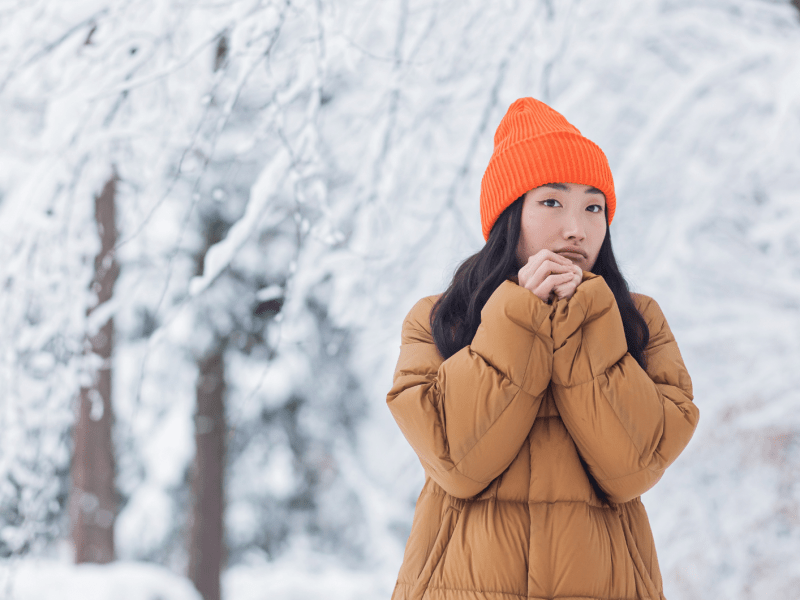 Eine junge Frau mit ernstem Blick wärmt ihre Hände, trägt eine orangefarbene Mütze und Daunenjacke vor verschneiten Bäumen.