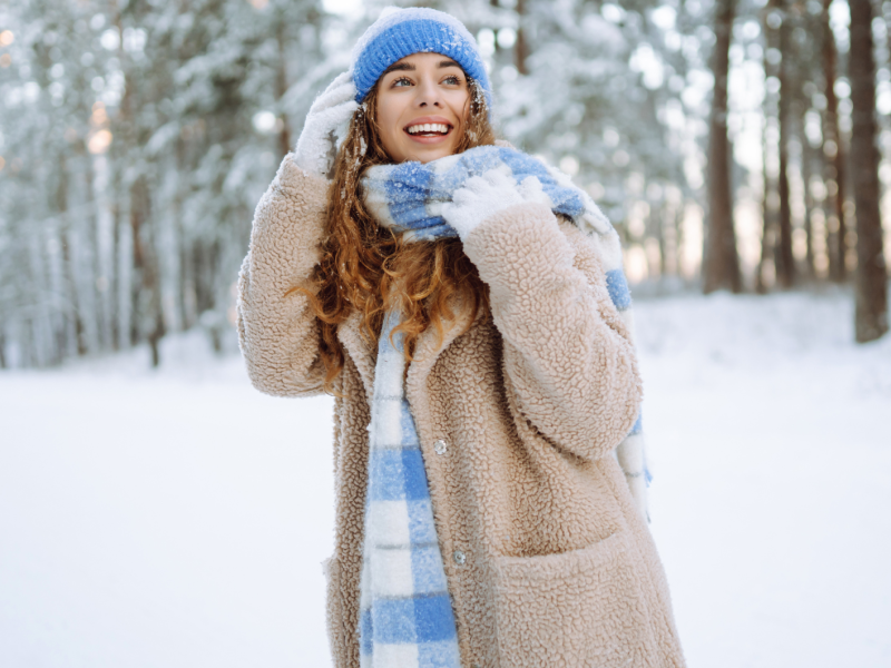 Junge Frau mit braunen Locken und blauer Mütze lacht fröhlich im verschneiten Winterwald, eingehüllt in einen beigen Mantel und karierten Schal.