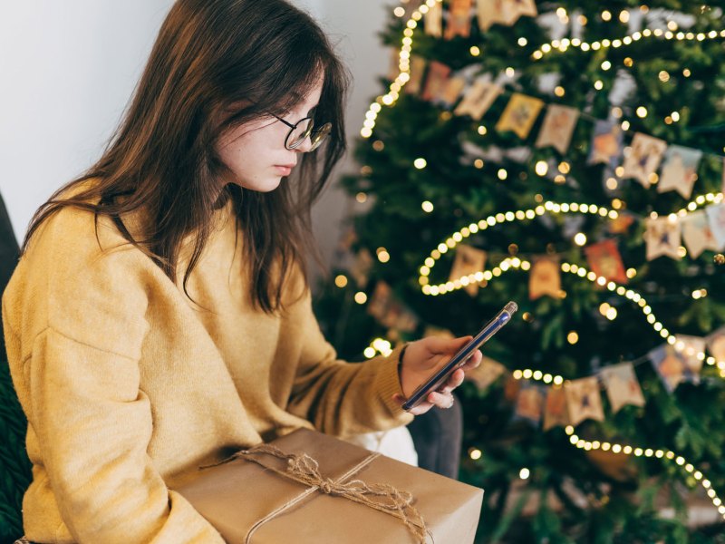 Teenagermädchen sitzt mit Geschenk auf dem Schoß vorm Weihnachtsbaum und schaut in ihr Handy.