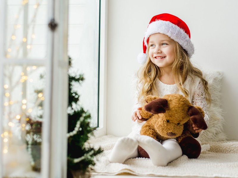 Mädchen, ca. 5 Jahre alt, sitzt mit einer Weihnachtsmannmütze am Fenster und schaut neugierig raus.