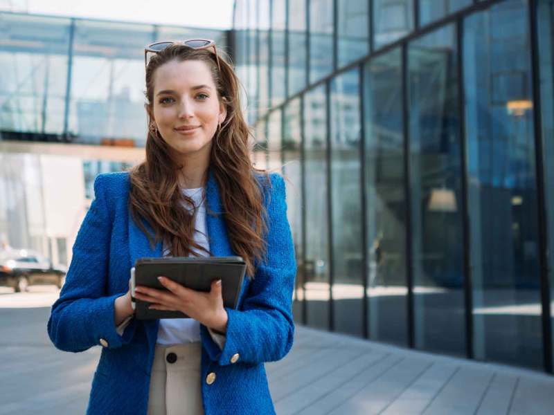 Frau mit braunen Haaren und einem blauen Blazer steht vor einem Bürogebäude und hält ein Tablet in den Händen.