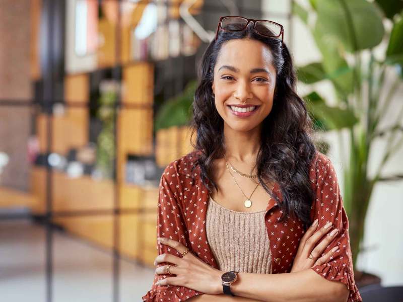 Junge Frau mit dunklen langen welligen Haaren steht lächelnd mit verschränkten Armen im Büro.