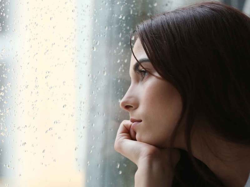 Eine dunkelhaarige Frau guckt im Januar traurig aus dem Fenster, während es draußen regnet.