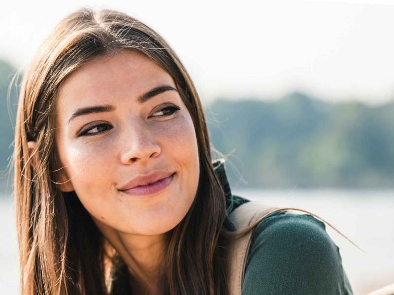 Junge Frau mit glatte dunklen Haaren sitzt draußen am Wasser und blickt freundlich zur Seite.