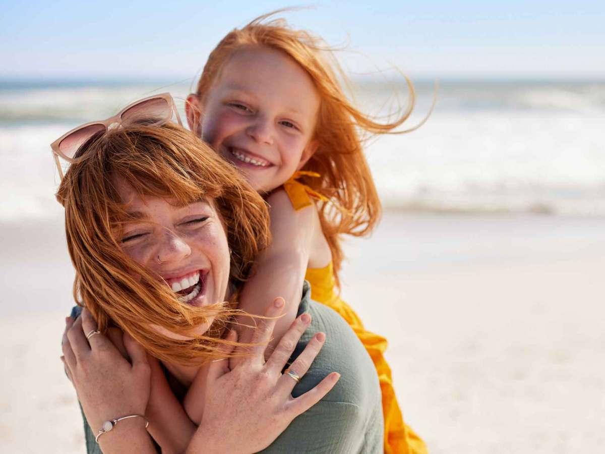 Eine lachende rothaarige Frau tr&auml;gt ein rothaariges M&auml;dchen in gelbem Kleid am sonnigen Strand, beide wirken ausgelassen.