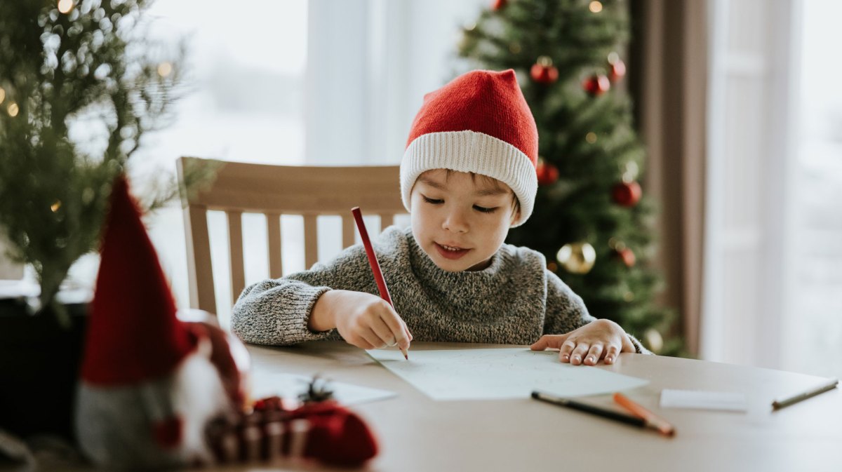 Kind sitzt am Tisch, trägt eine Weihnachtsmannmütze und malt einen Wunschzettel.
