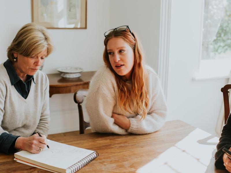 Drei Personen sitzen an einem Holztisch in einem hellen Raum, eine ältere Frau zeichnet, während eine junge Frau mit roten Haaren spricht.