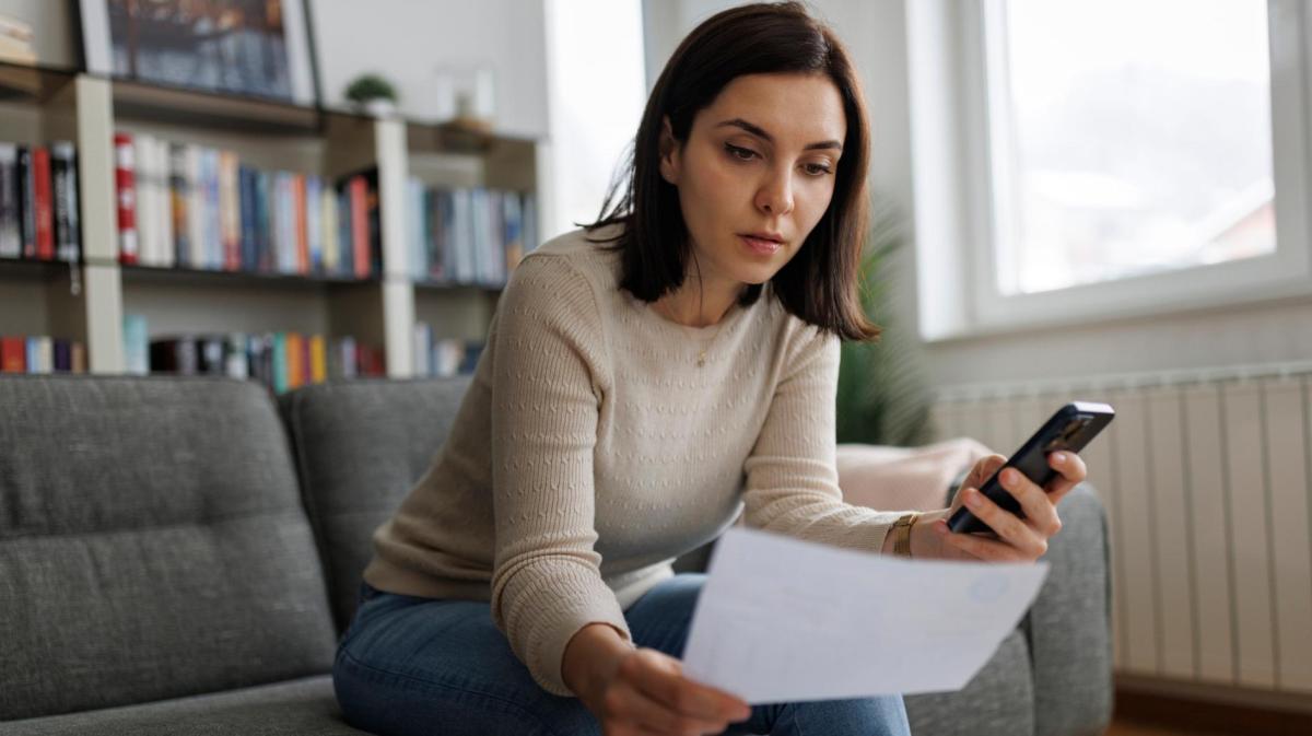 Eine junge Frau mit schulterlangem braunem Haar sitzt nachdenklich auf einem grauen Sofa, hält ein Dokument und schaut auf ihr Smartphone.