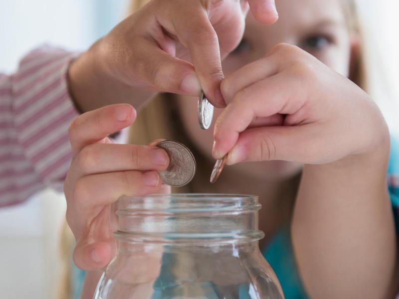Drei Kinderhände werfen konzentriert Münzen in ein großes, leeres Einmachglas vor unscharfem, hellem Hintergrund.