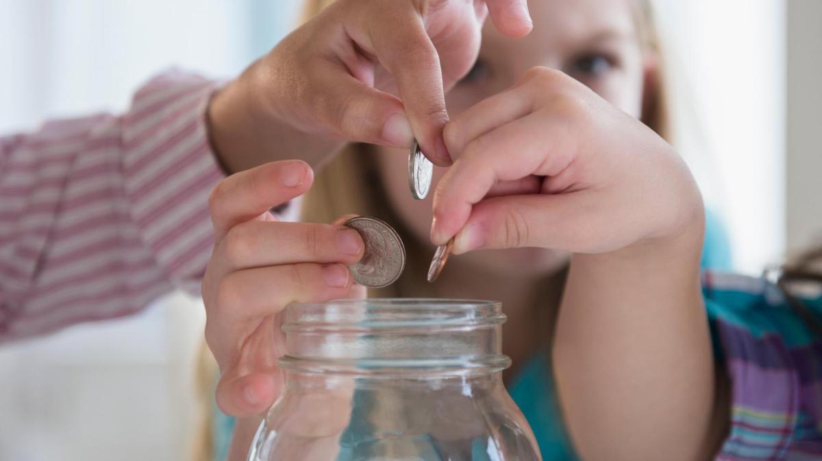 Drei Kinderhände werfen konzentriert Münzen in ein großes, leeres Einmachglas vor unscharfem, hellem Hintergrund.