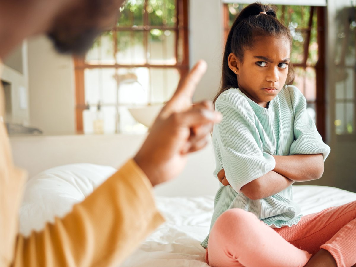 Wütendes Mädchen sitzt auf ihrem Bett, verschränkt die Arme und schaut ihren Vater böse an.