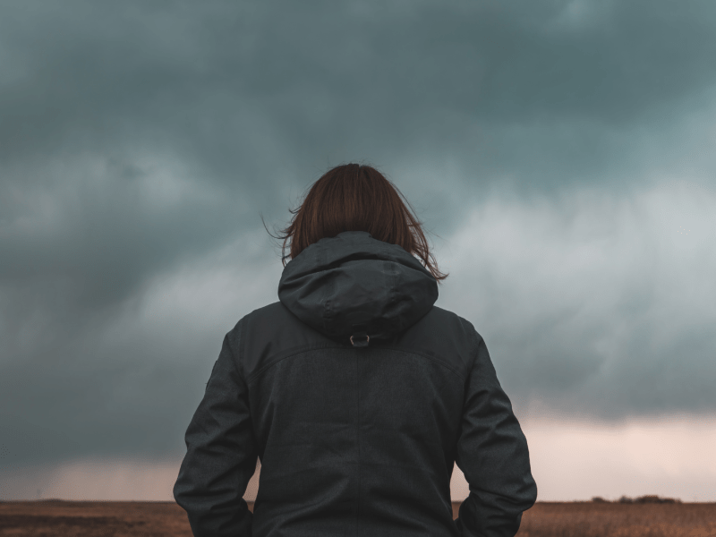 Frau steht mit dem Rücken zur Kamera vor einem düsteren Wolkenhimmel