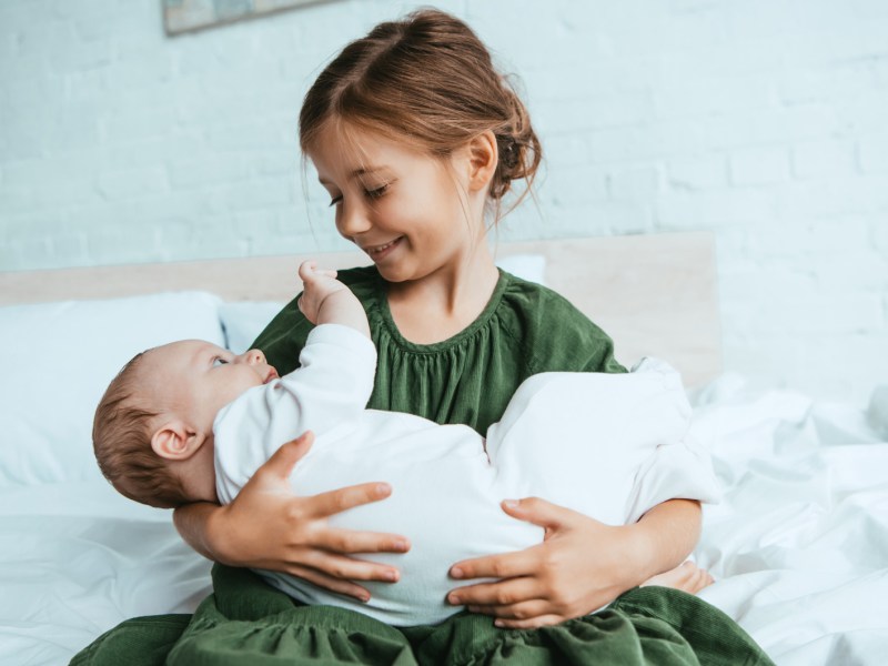 Glückliches Mädchen hält ihre kleine Baby-Schwester im Arm, während sie auf einem weißen Bett sitzt.