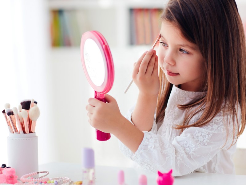 Kleines Mädchen, ca. 6 bis 8 Jahre, sitzt mit einem Handspiegel am Tisch und trägt mit einem Make-up-Pinsel Augen-Make-up auf.