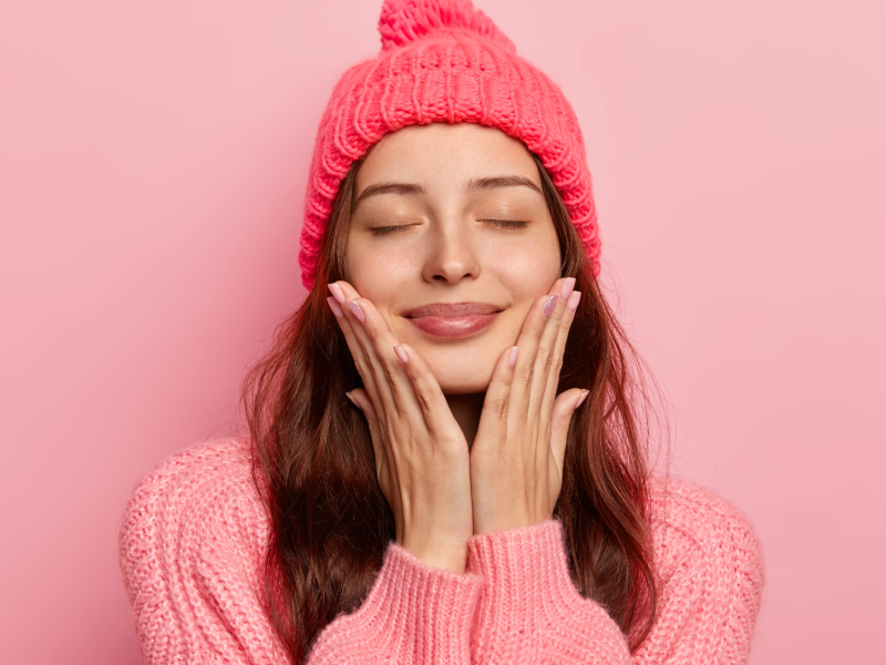 Junge Frau mit geschlossenen Augen lächelt entspannt, trägt eine pinke Strickmütze und Pullover vor rosa Hintergrund.
