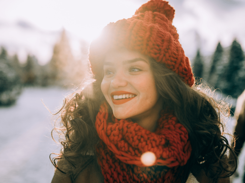 Junge Frau mit gelockten, dunkelbraunen Haaren und einer roten Mütze und einem roten Schal, die lächelnd zur Seite schaut, mit der Sonne im Hintergrund.