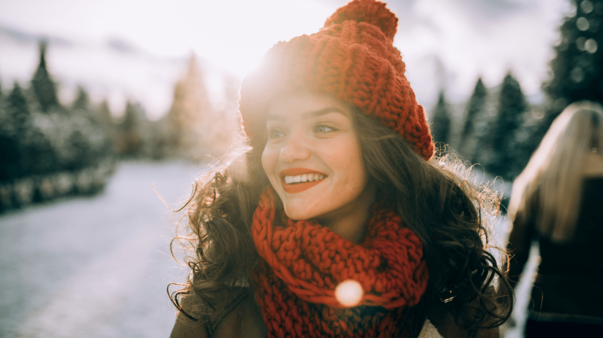 Junge Frau mit gelockten, dunkelbraunen Haaren und einer roten Mütze und einem roten Schal, die lächelnd zur Seite schaut, mit der Sonne im Hintergrund.