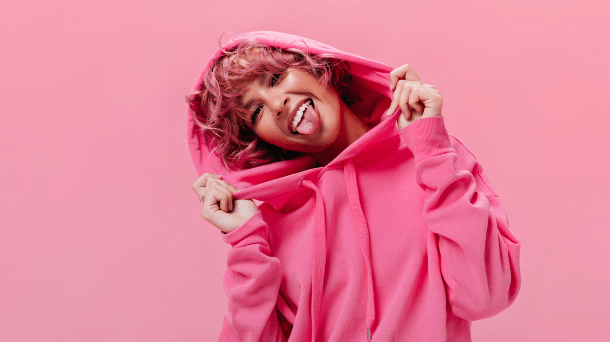 Frau mit pinken Kapuzenpulli, vor einer punken Wand mit pinken Haaren, die ihre Zunge herausstreckt und lächelt.