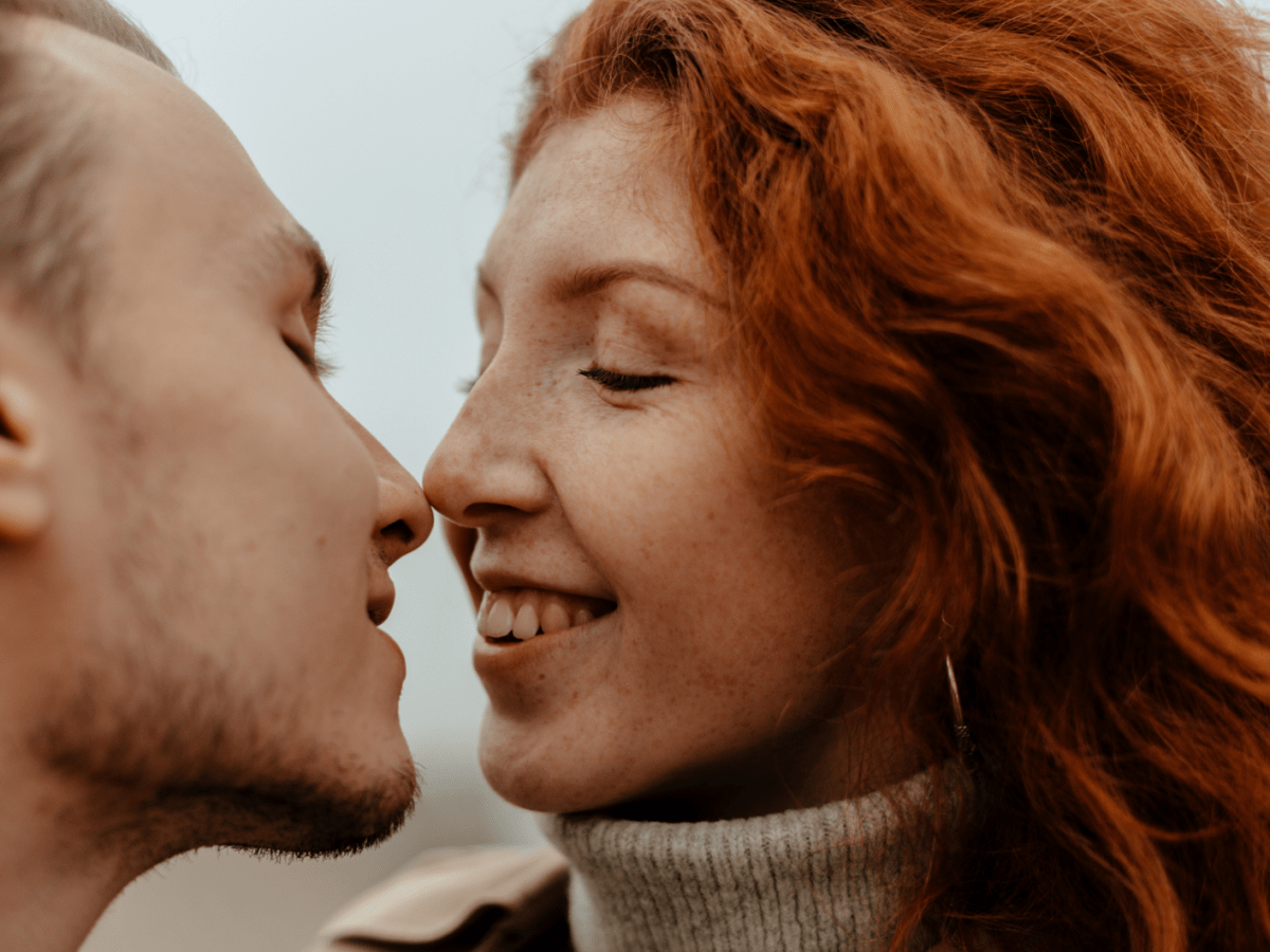 Frau mit roten Haaren k&uuml;sst einen Mann mit dunklen, kurzen Haaren und l&auml;chelt.