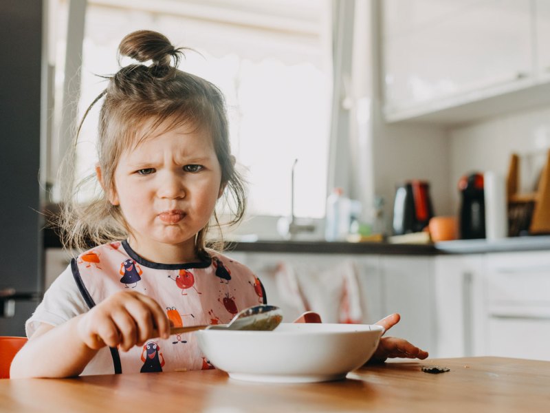 Süßes, kleines Mädchen, das ein witziges Gesicht zieht beim Essen.