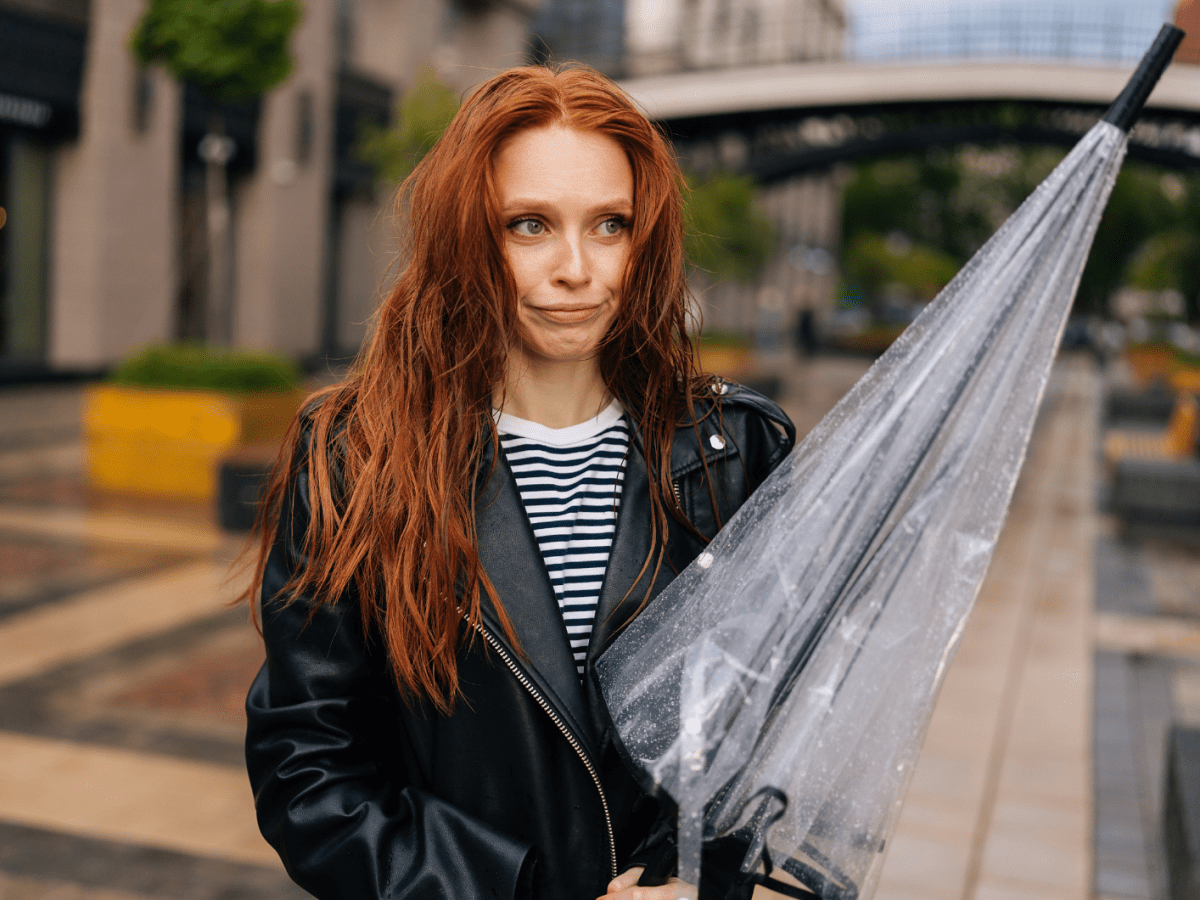 Frau mit nassen und durchwuschelten Haaren, die einen Regenschirm in der Hand hält und schmollend zur Seite schaut.