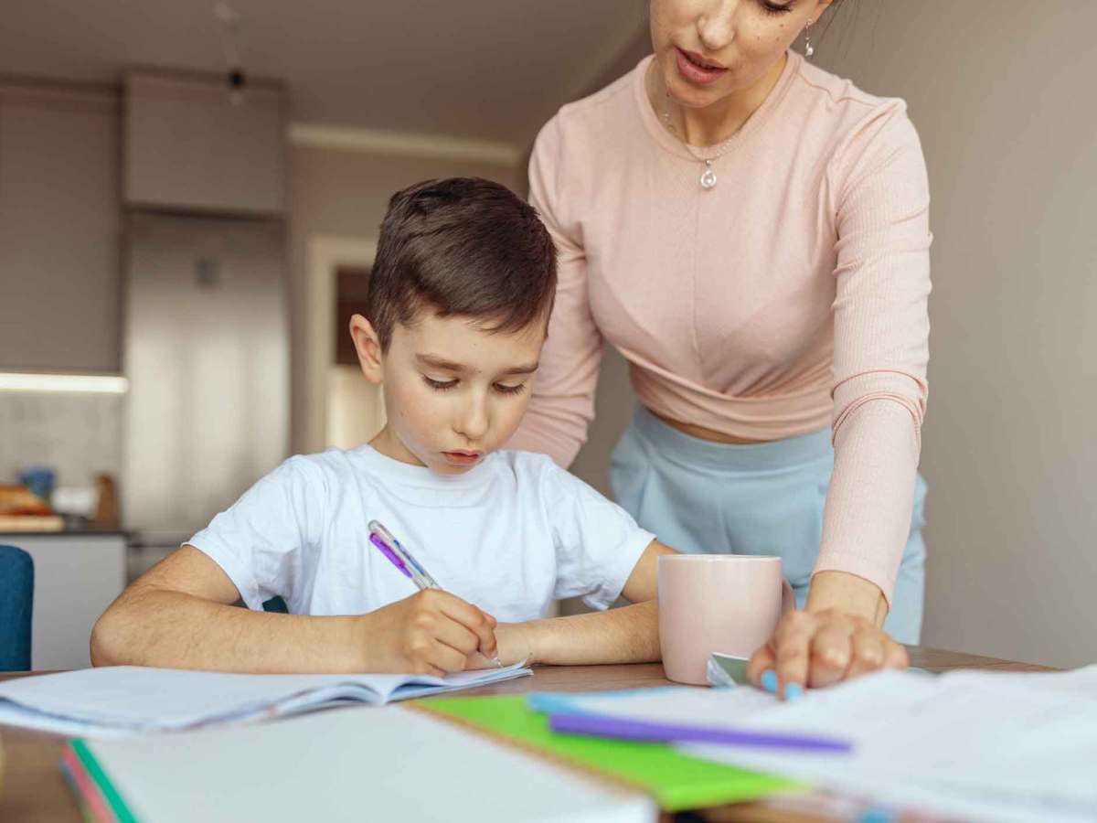 Ein Grundschulkind arbeitet mit Stift an den Hausaufgaben, neben ihm steht eine Frau und zeigt auf ein Blatt.