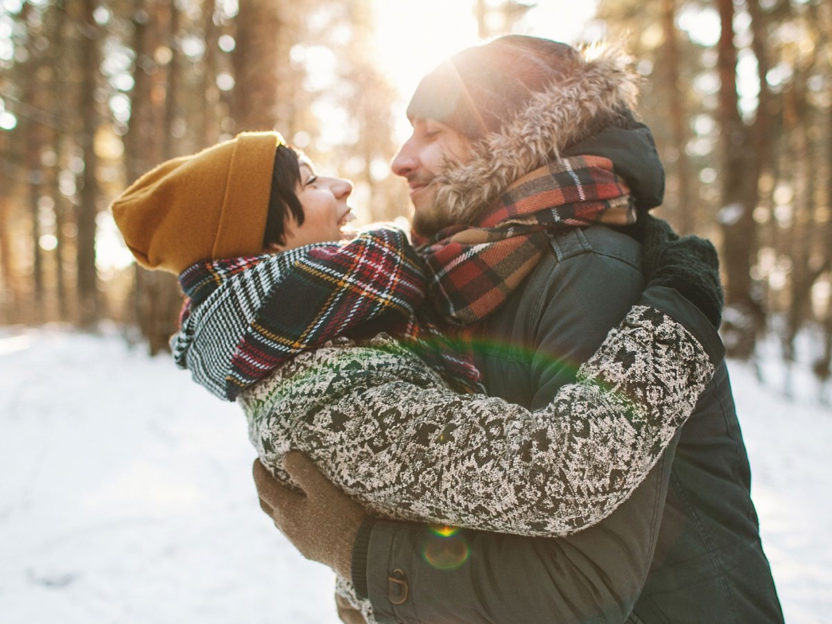 Mann und Frau in Winterbekleidung lachen und umarmen sich in verschneitem Wald