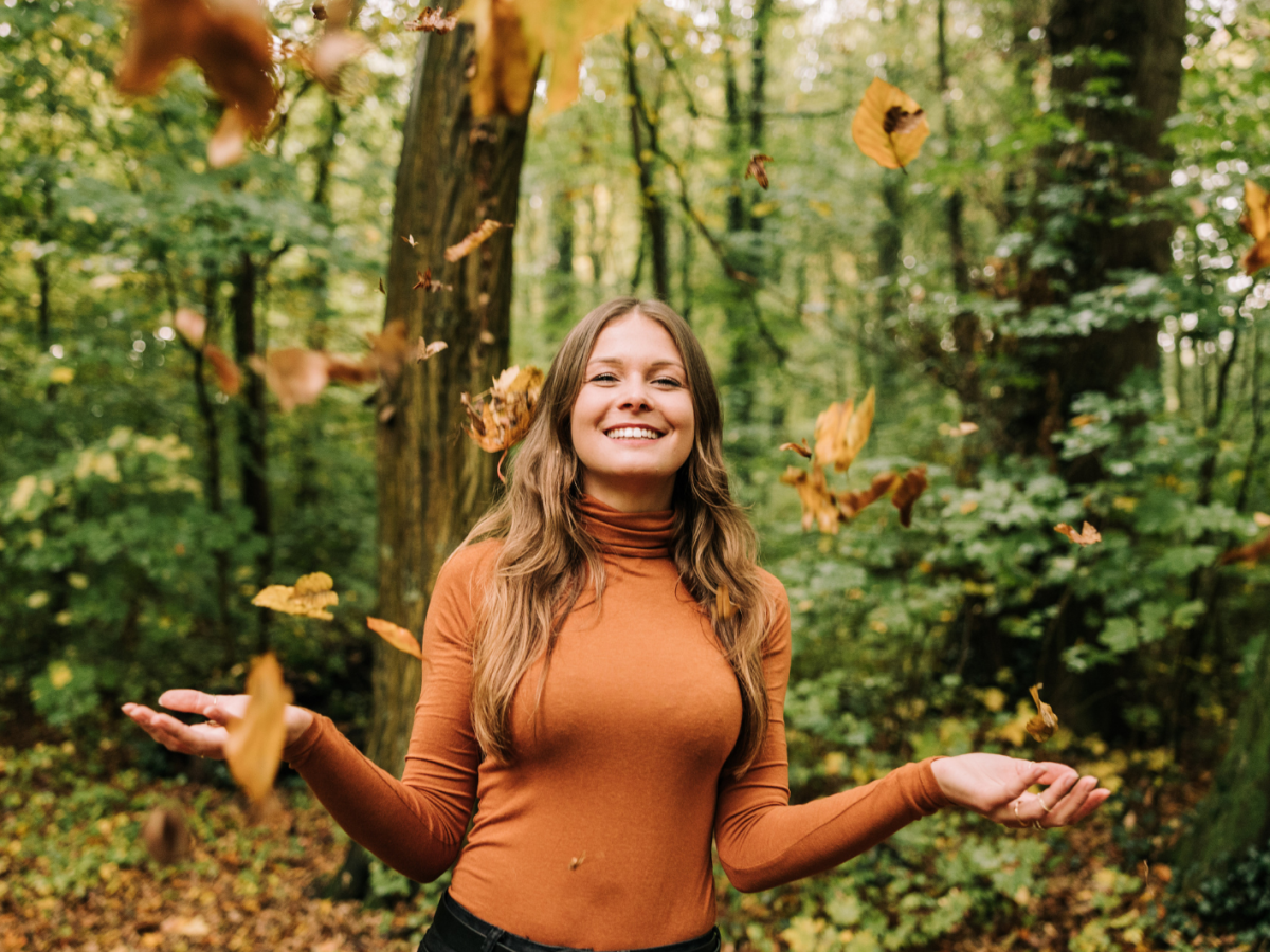 Frau im Wald, die lacht und Blätter in die Luft wirft.
