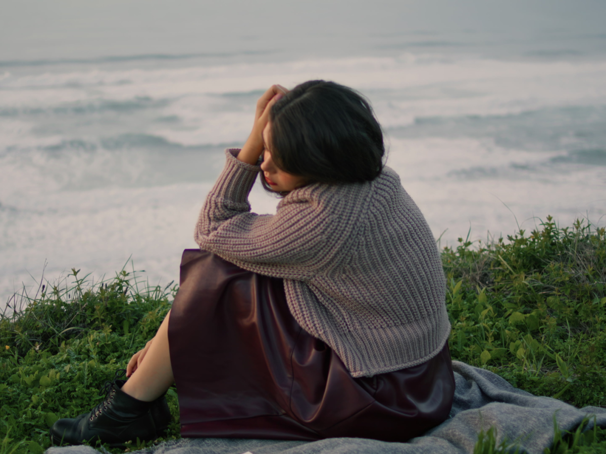 Nachdenkliche Frau mit kurzem dunklem Haar, Strickpullover und Lederrock sitzt auf einer Decke vor einer welligen Meereskulisse.