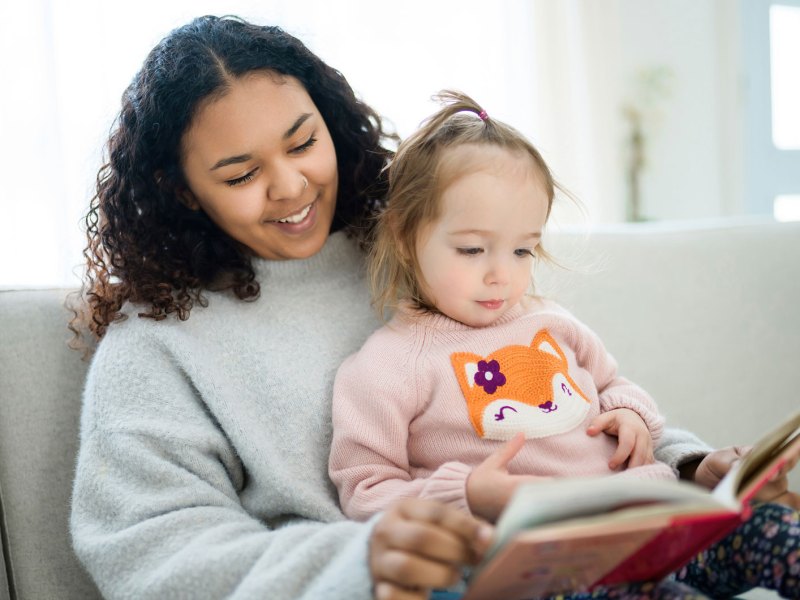 Mädchen im Teenageralter passt auf ein Mädchen von ca. 4 Jahren auf. Sie lesen ein Buch zusammen.