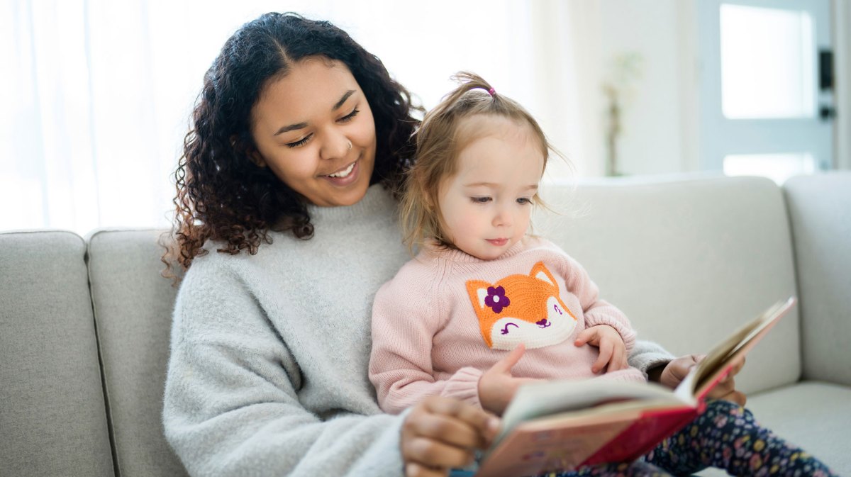Mädchen im Teenageralter passt auf ein Mädchen von ca. 4 Jahren auf. Sie lesen ein Buch zusammen.