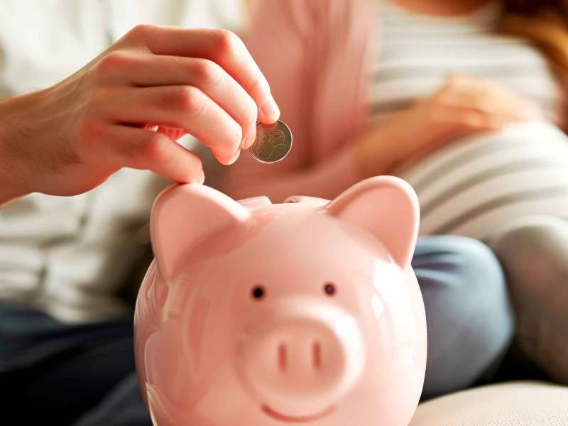 Mann wirft eine Münze in ein rosa Sparschwein. Im Hintergrund sitzt eine schwangere Frau.