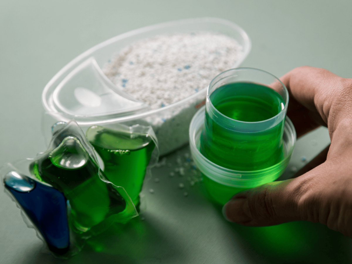 Person stellt Waschpulver, Flüssigwaschmittel & Wäsche-Pods auf eine Ablage