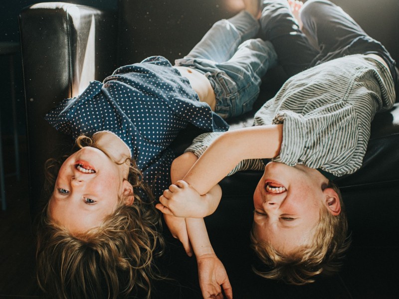 Zwei Kinder liegen Kopf nach unten auf einem dunklen Sofa