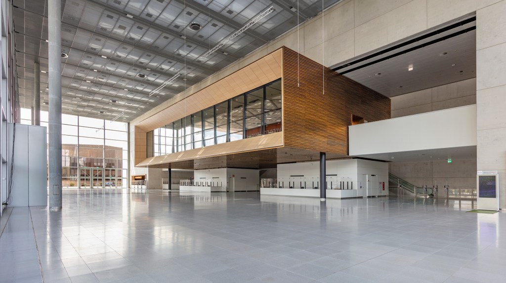 Moderne, lichtdurchflutete Empfangshalle mit Holz- und Betonelementen, großen Fenstern und Rolltreppen im Hintergrund.