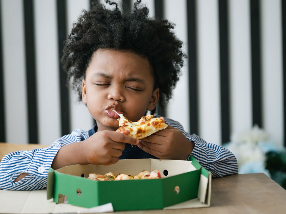Gl&uuml;cklihces Kind bei&szlig;t genussvoll und mit geschlossenen Augen in ein St&uuml;ck Pizza. Vor ihm auf dem Tisch steht der Pizzakarton.