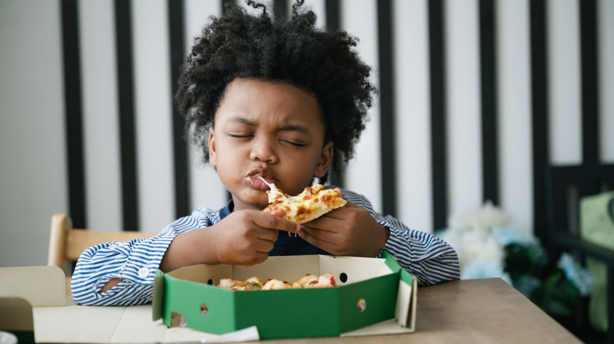 Glücklihces Kind beißt genussvoll und mit geschlossenen Augen in ein Stück Pizza. Vor ihm auf dem Tisch steht der Pizzakarton.