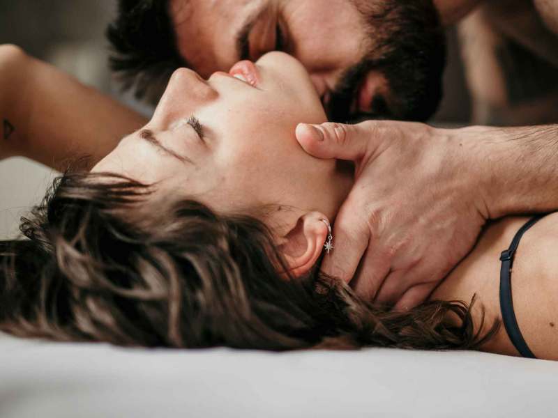 Eine Frau liegt im Bett und ihr Partner küsst sie leidenschaftlich am Hals, während sie die Augen geschlossen hat.