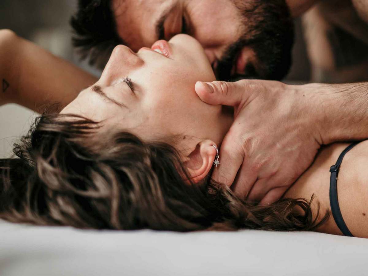 Eine Frau liegt im Bett und ihr Partner küsst sie leidenschaftlich am Hals, während sie die Augen geschlossen hat.