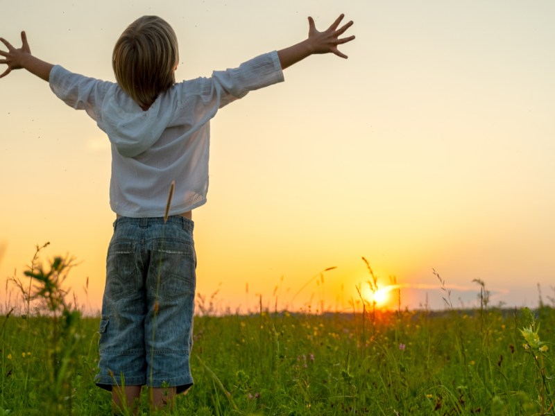 Glückliches Kind steht auf einem Feld bei Sonnenuntergang und umarmt die Welt.