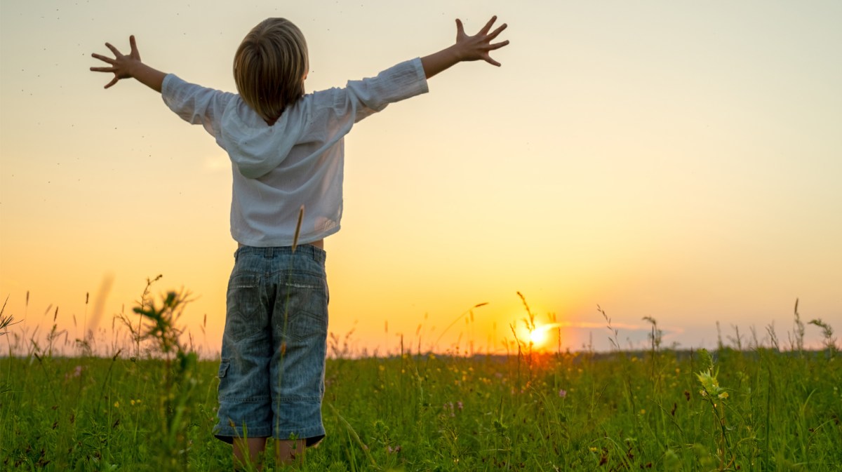 Glückliches Kind steht auf einem Feld bei Sonnenuntergang und umarmt die Welt.