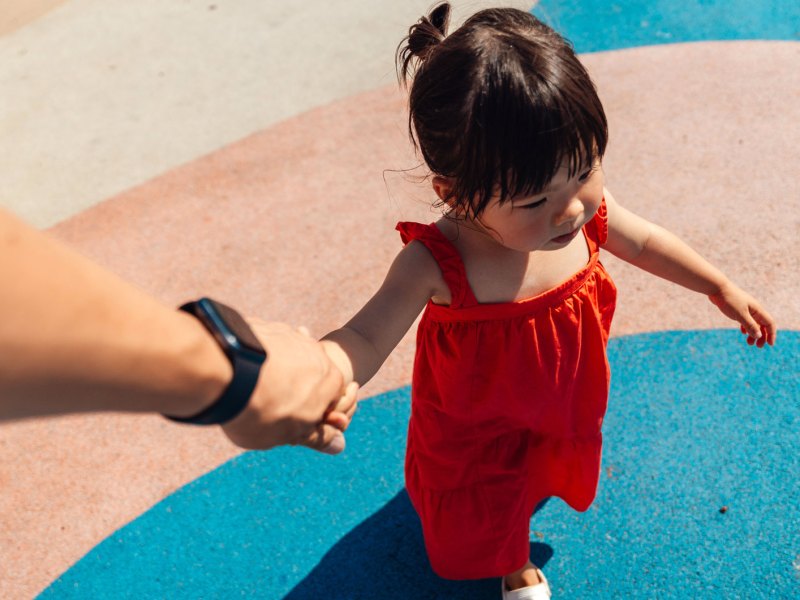 Mädchen in rotem Kleid an Hand von Erwachsenem als Symbolbild für Helikopter-Eltern
