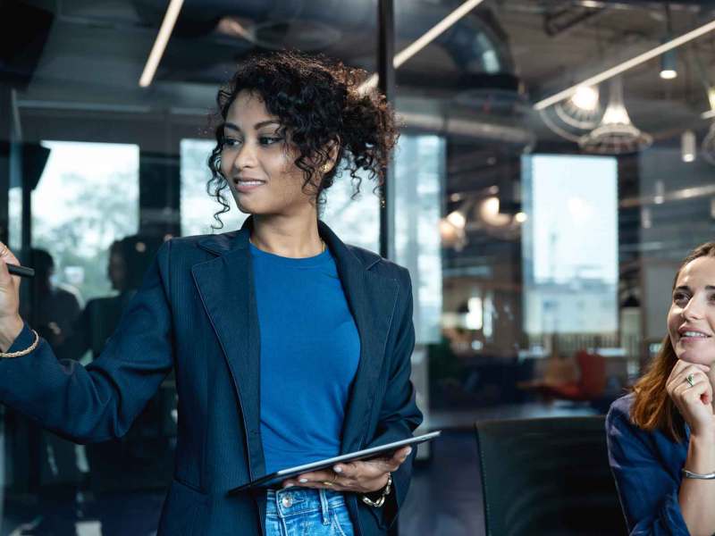 Eine junge Frau mit dunklen Locken und blauem Blazer schreibt lächelnd auf eine Glaswand, während eine Kollegin sie bewundernd ansieht.