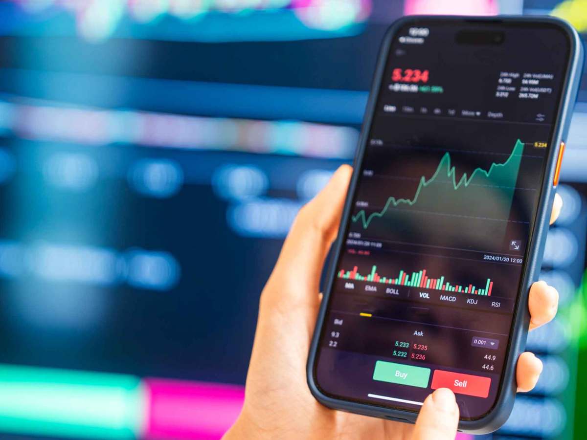 Eine Hand hält ein Smartphone mit Börsen-App, das steigende Aktienkurse zeigt, im Hintergrund unscharfe Börsendiagramme.