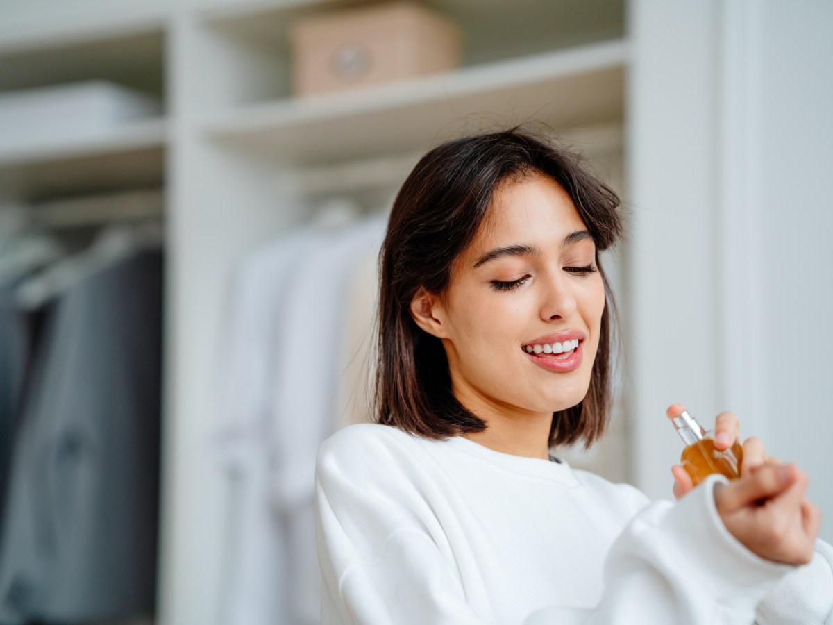 Frau in weißem Pullover sprüht sich Parfum auf und lächelt