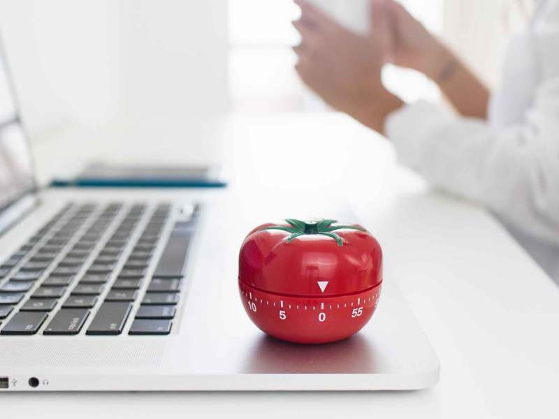 Eine Küchenuhr in Form einer Tomate steht auf einem Notebook. Im Hintergrund sitzt eine Frau mit einem Kaffee.