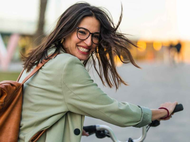 Frau mit dunklen Haaren und Rucksack schiebt lachend ihr Fahrrad.