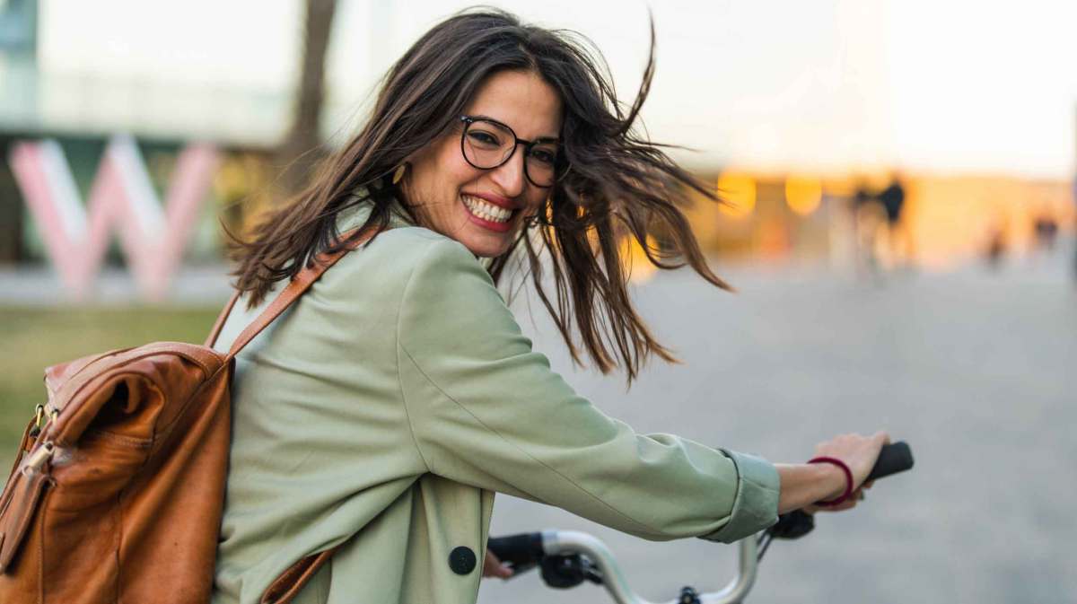 Frau mit dunklen Haaren und Rucksack schiebt lachend ihr Fahrrad.