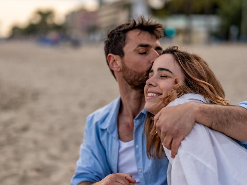Ein Mann mit Dreitagebart in offenem Jeanshemd küsst eine lachende Frau mit welligen, braunen Haaren auf die Stirn am Sandstrand.